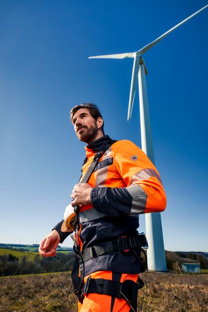 A man wears Multinorm protective clothing in orange and black. A wind turbine can be seen in the background.
